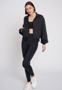 Giacca sportiva nera con zip, texture morbida e polsini a costine. Indossata sopra un top corto nero abbinato e leggings, abbinata a sneakers bianche.