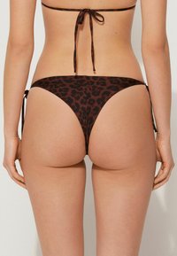 Bikini kalhotky s leopardím vzorem, vyrobené z měkké tkaniny. Obsahují postranní šňůrky, nízký pas a design s minimálním zakrytím v hnědých a černých odstínech.