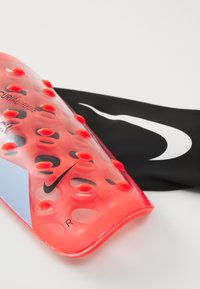 Protège-tibias Nike rouges avec des reliefs en forme de petits ronds et logo swoosh noir, reposant sur un tissu noir avec un grand swoosh Nike blanc.
