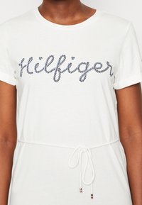 White T-shirt with short sleeves, featuring a navy rope-style embroidered logo. Elastic drawstring waist with metal accents. Soft cotton material.