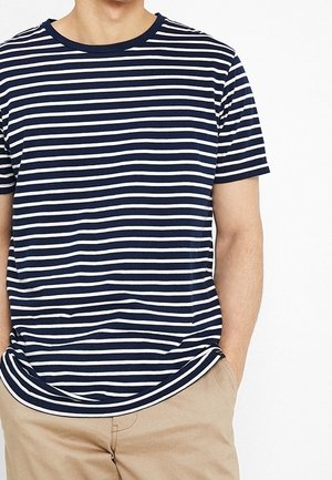 Mężczyzna ubrany w granatowo-biały, poziomo paskowany t-shirt z krótkim rękawem i beżowe spodnie, ręce w kieszeniach, na białym tle.