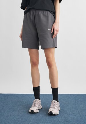 Osoba oblečená v tmavosivých šortkách so sťahovacou šnúrkou, čiernych ponožkách a svetlosivých teniskách stojí na modrom koberci pred jednoduchou stenou.