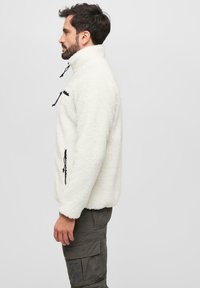 Valge fleece pullover kõrgema kraega, kahe tõmblukuga tasku ja mustade nööridega. Tekstuuriga kangas, millel on pehme ja kurruline välimus.
