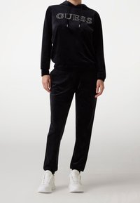 Zwarte velours trainingspak met een pullover hoodie waarop "GUESS" in strasstenen staat, gecombineerd met taps toelopende broek en witte sneakers.