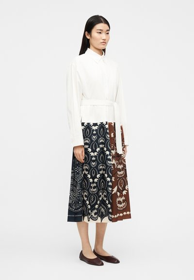 Γυναίκα στέκεται φορώντας λευκό  πουκάμισο με μακριά μανίκια και ζώνη, φούστα μέση μήκος με πλισέ σχέδιο σε ναυτικό μπλε και καφέ, και σκούρα καφέ επίπεδα παπούτσια.