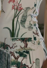 Bloemmotief jurk met groene bladeren en bruine bomen, gemaakt van zachte stof. Bevat een klein zakje en decoratieve hardware.