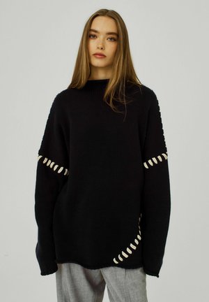 Pull en tricot noir avec une coupe ample, doté d'accents de couture blanche le long des manches. Le design présente une texture côtelée et un col rond.