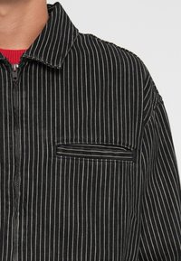 Chaqueta de rayas en blanco y negro con cremallera frontal, cuello y un bolsillo en el pecho. Presenta una tela texturizada y un patrón vertical prominente.