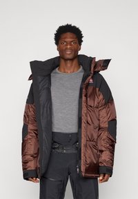 Giacca puffa con una combinazione di colori lucidi in marrone e nero, dotata di un colletto alto, chiusura con zip e cappuccio regolabile. Dettagli in tessuto testurizzato.