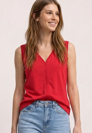 Uśmiechnięta kobieta z długimi włosami, ubrana w czerwoną bluzkę bez rękawów i jasnoniebieskie dżinsy, patrząca w swoją prawą stronę na jednolitym tle.