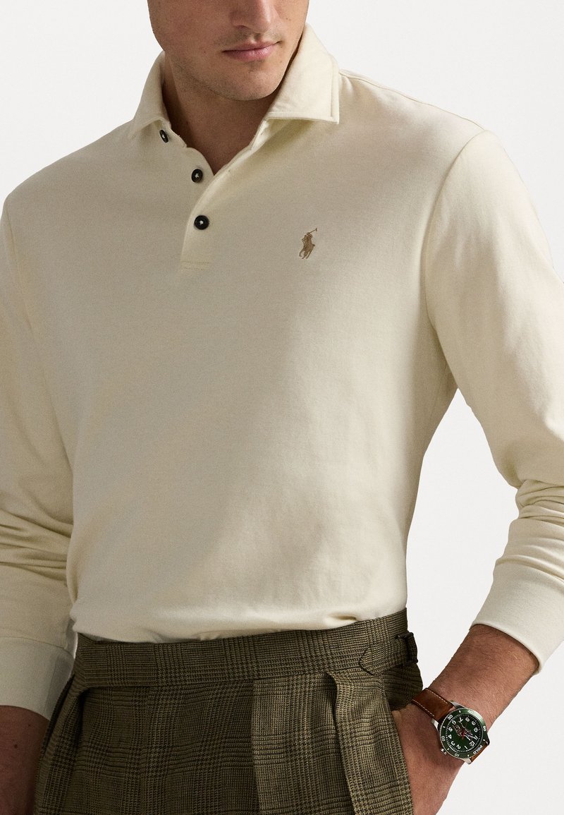 Beige polo shirt met lange mouwen, een knoopsluiting bij de kraag en een klein geborduurd logo. Gecombineerd met gestreepte broeken en een leren bruine horloge.