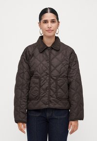 Woman with dark hair in gold hoop earrings wearing a dark brown quilted jacket with patch pockets and button closure, standing against white background.