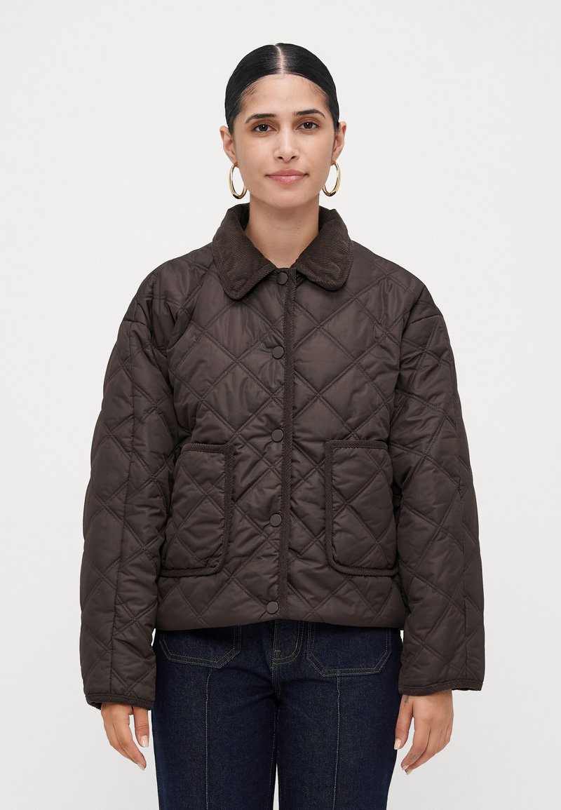 Woman with dark hair in gold hoop earrings wearing a dark brown quilted jacket with patch pockets and button closure, standing against white background.