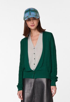 Grüne Strickjacke mit tiefem V-Ausschnitt, langen Ärmeln und einer Vorderknopf. Getragen über einem hellgrauen, knöpfbaren Oberteil, kombiniert mit einer blauen karrierten Kappe.