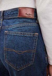 Jeans de ganga azul escuro com um logo "Pepe Jeans" em um retalho de couro castanho. Costura laranja em contraste visível nos bolsos e costuras.