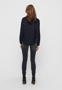 Haut texturé à manches longues de couleur marine avec un ourlet arrondi, associé à un jean ajusté gris et des bottes à talons noires. Vue arrière, fond uni.