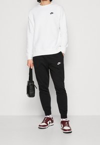 Osoba v bílé mikině Nike, černých teplácích Nike, maroon a bílých teniskách Nike, držící černou koženou tašku za popruh.