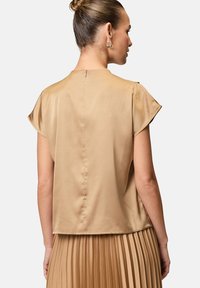 Top corto a maniche corte in satin beige con collo alto; presenta una zip centrale nella parte posteriore e una texture liscia, abbinato a una gonna plissettata.