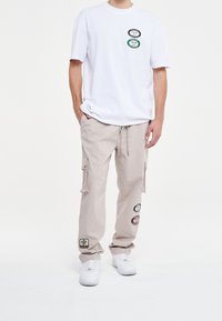 Witte T-shirt met korte mouwen en drie ronde patches, gecombineerd met beige cargobroek met zijzakken en gestikte branding, gedragen met witte sneakers.