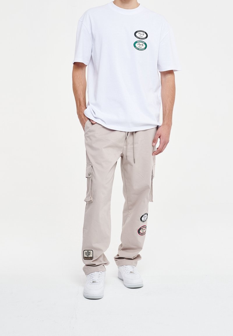Witte T-shirt met korte mouwen en drie ronde patches, gecombineerd met beige cargobroek met zijzakken en gestikte branding, gedragen met witte sneakers.
