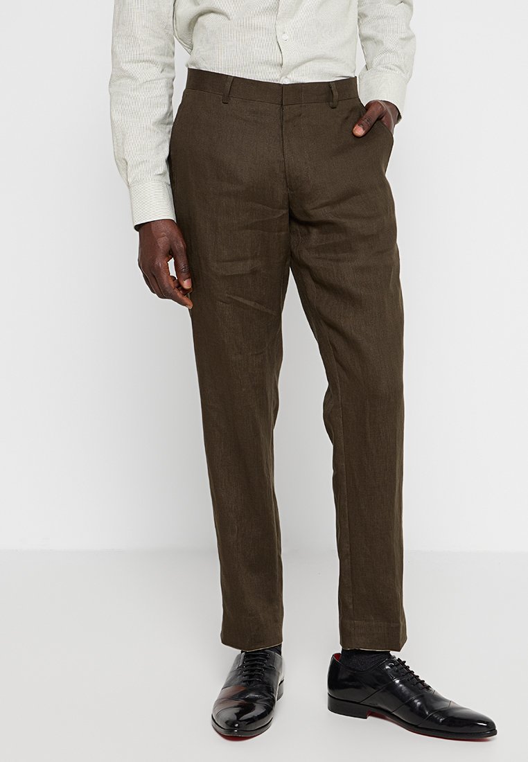 Mann in dunkelbraunen Maßanzughosen, mit einem hellen gemusterten Hemd und schwarzen polierten Schnürschuhen, steht mit einer Hand in der Tasche.