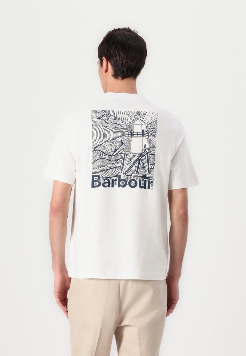 Homme portant un T-shirt blanc avec un phare en art linéaire noir et le texte "Barbour" dans le dos, assorti à un pantalon beige.
