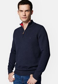 Granatowy sweter rozpinany wykonany z dzianiny, z wysokim kołnierzem, ściągaczami przy mankietach oraz w kratkę w środku. Model nosi dżinsy.
