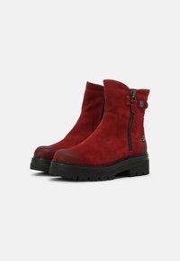 Botas de tobillo de ante rojo con suela de goma negra, cremallera lateral y un botón decorativo; punta redondeada, textura suave y tacón bajo tipo bloque.