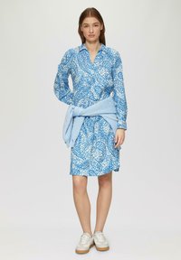 Blaumuster Kleid mit langen Ärmeln und Knopfkragen, kombiniert mit einem hellblauen Strickpullover, der um die Taille gebunden ist. Weiße Sneaker.