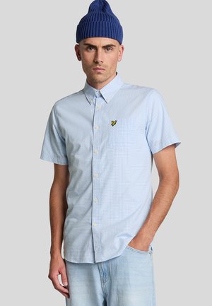 Chemise à manches courtes en vichy bleu clair avec un col boutonné et une poche poitrine arborant un petit logo jaune. Portée avec un denim.
