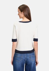Witte trui met korte mouwen en marineblauwe afwerking, met een gestructureerde stof en een getailleerd ontwerp, gecombineerd met blauwe jeans die zichtbaar zijn op de taille.