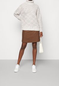 Gehaakt coltrui in lichtcrème met multikleurige stippen, gecombineerd met een nauwsluitende bruine rok en witte sneakers. Houdt een kleine witte tas vast.