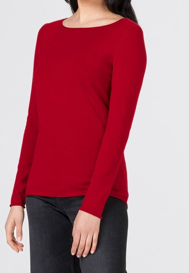 Femme portant un haut rouge ajusté à manches longues avec un large col rond et un jean noir, debout devant un fond gris uni.