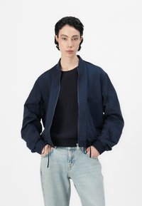 Marineblå bomberjakke med glidelås foran, ribbestrikkede mansjetter og justerbar snor i nederkanten, kombinert med lyseblå denimjeans og en mørk genser.