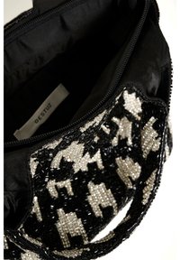 Černá kabelka zdobená korálky s dekorativním vzorem bílých a stříbrných motivů. Má zipové uzavírání a černou látkovou podšívku. Uvnitř je viditelná štítek.