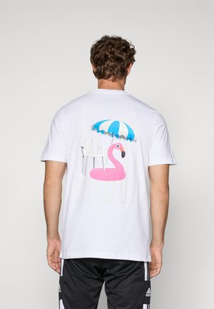 Man die een wit T-shirt draagt met een afbeelding van een roze flamingo-zwembaddrijf, een witte plastic stoel en een blauw-witte strandparasol op de achtergrond.