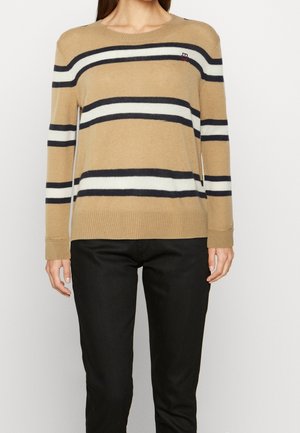 Femme portant un pull beige à rayures horizontales noires et blanches associé à un pantalon noir, debout devant un fond blanc.