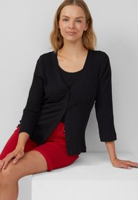 Zwarte geribbelde cardigan met V-hals, driekwartmouwen en knopen aan de voorkant, gecombineerd met rode shorts op een witte ondergrond.