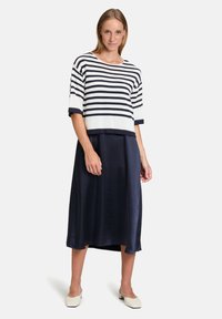 Frau in einem gestreiften Kurzarmoberteil in Weiß und Marineblau, einem marineblauen Midirock und weißen flachen Schuhen, die vor einem schlichten Hintergrund steht.