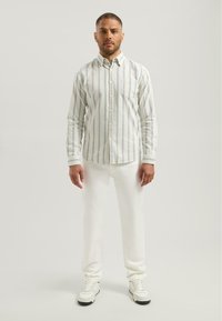 Mężczyzna stoi przodem, ubrany w białe spodnie, białe trampki i białą koszulę z guzikami w pionowe jasnoniebieskie paski.