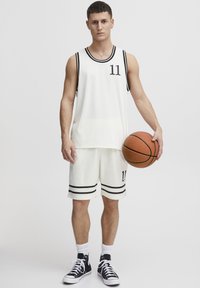 Hvid basketballtrøje og -shorts med mesh-stof, kontrasterende sorte striber og nummer 11. Modellen holder en basketball.