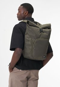Khaki roll-top backpack made of durable fabric, featuring side pockets, adjustable straps, and a front zip pocket for secure storage.