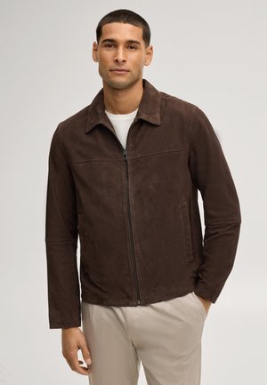 Uomo con capelli corti indossa una giacca marrone con cerniera sopra una camicia bianca e pantaloni beige, in piedi con una mano in tasca su sfondo grigio.