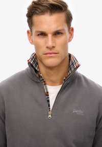 Suéter gris con cremallera y un cuello interior de cuadros. Presenta una cremallera plateada y el logo de SuperDry bordado en gris en el pecho.
