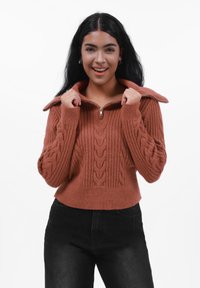 Kabelstrikket, rustfarget genser med glidelås og høy krage, og ribbestruktur; modellen har den på med svarte høytlivsjeans.