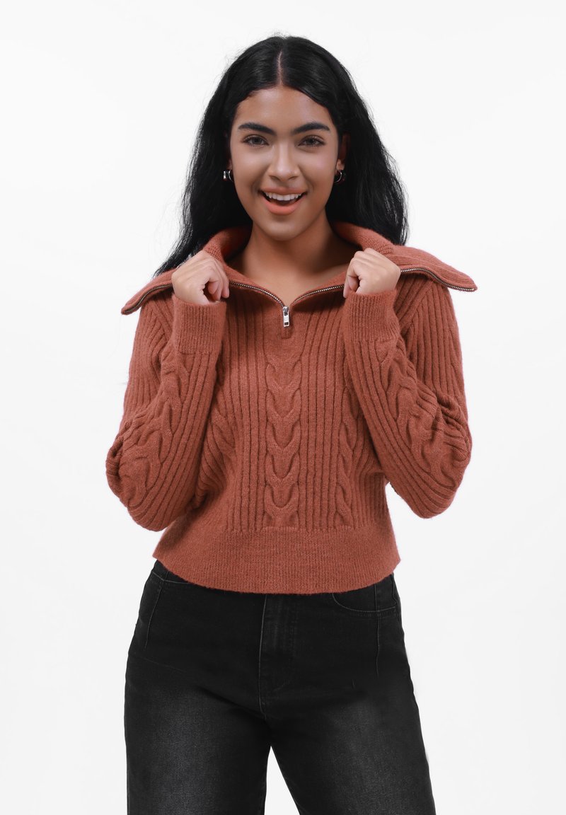 Kabelstrikket, rustfarget genser med glidelås og høy krage, og ribbestruktur; modellen har den på med svarte høytlivsjeans.