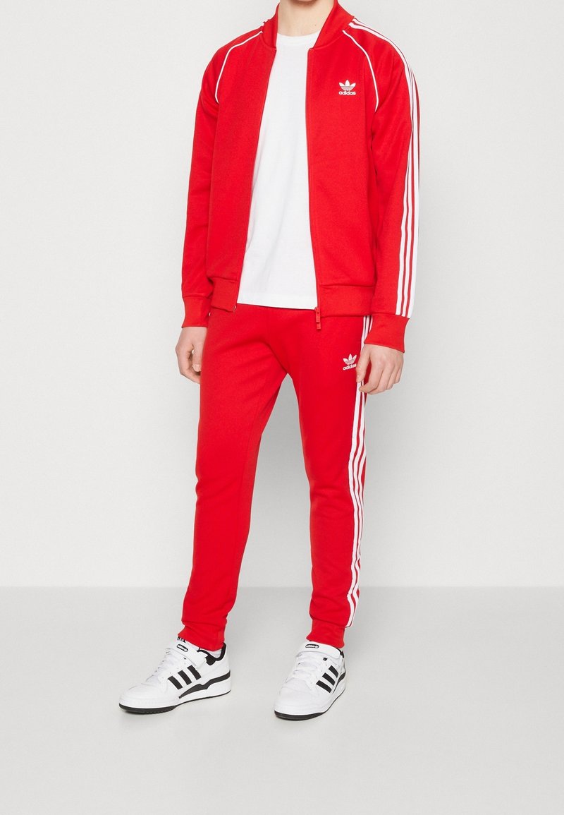 Röd träningsdräkt med vita ränder på ärmarna och byxorna, zippered jacka, kombinerad med en vit t-shirt och svartvita sneakers.