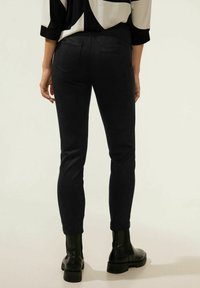 Pantaloni neri slim-fit con una texture liscia, dotati di tasche posteriori. Indossati con stivaletti neri alla caviglia e una blusa con fantasia nei toni del nero e bianco.
