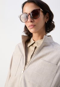 Giacca beige con zip dal finish testurizzato, dotata di colletto e due tasche anteriori. Grandi occhiali da sole tartaruga completano il look.