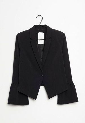 Blazer negro con cierre de un solo botón, puños acampanados y solapas con muescas colgado de una percha negra con una etiqueta de precio blanca.
